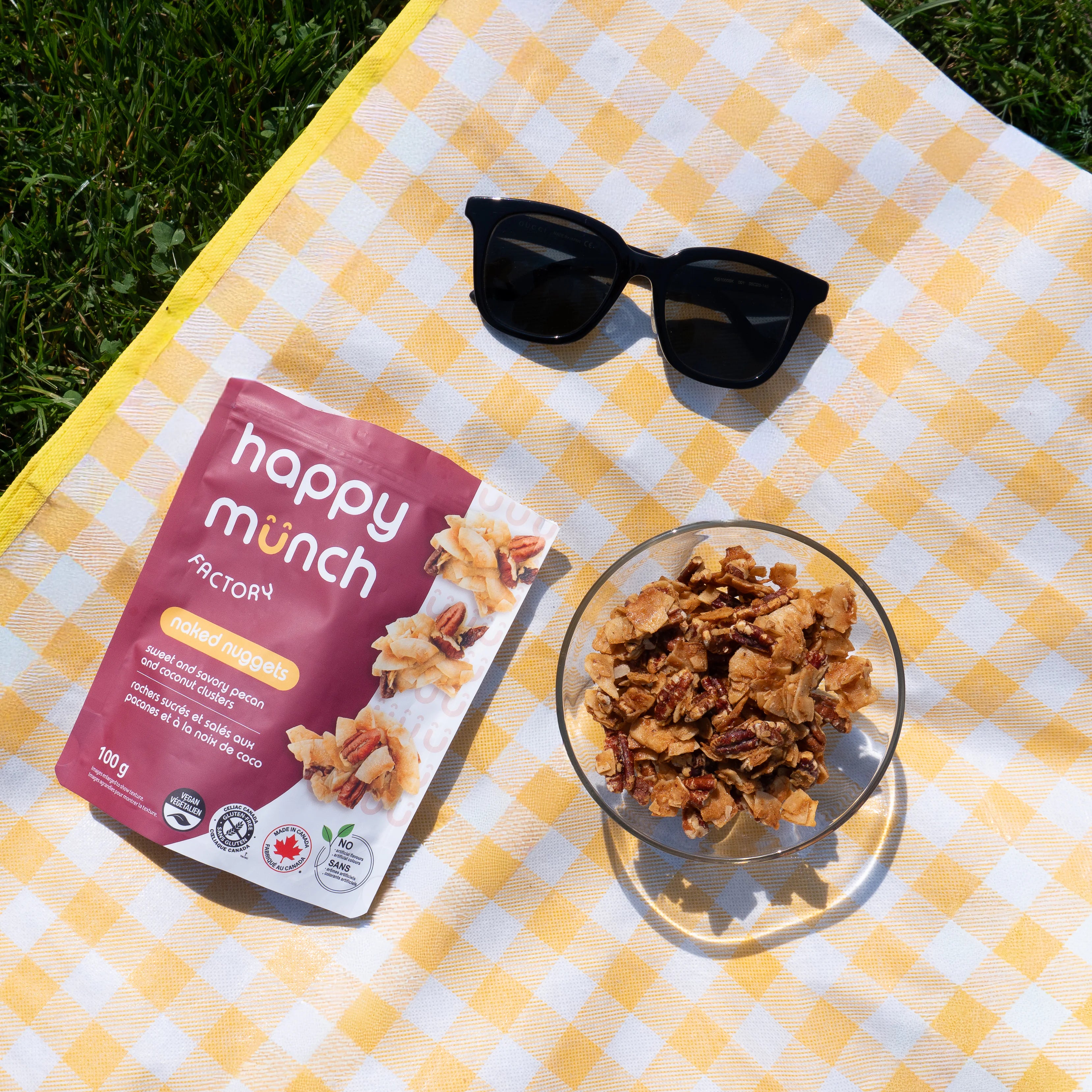 Happy Munch Factory Naked Nugget package with a bowl of Naked nugget coconut clusters on a yellow checkered blanket outdoors.