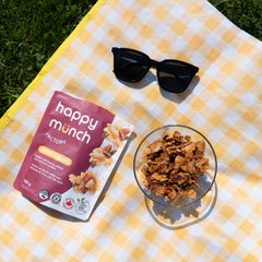 Happy Munch Factory Naked Nugget package with a bowl of Naked nugget coconut clusters on a yellow checkered blanket outdoors.