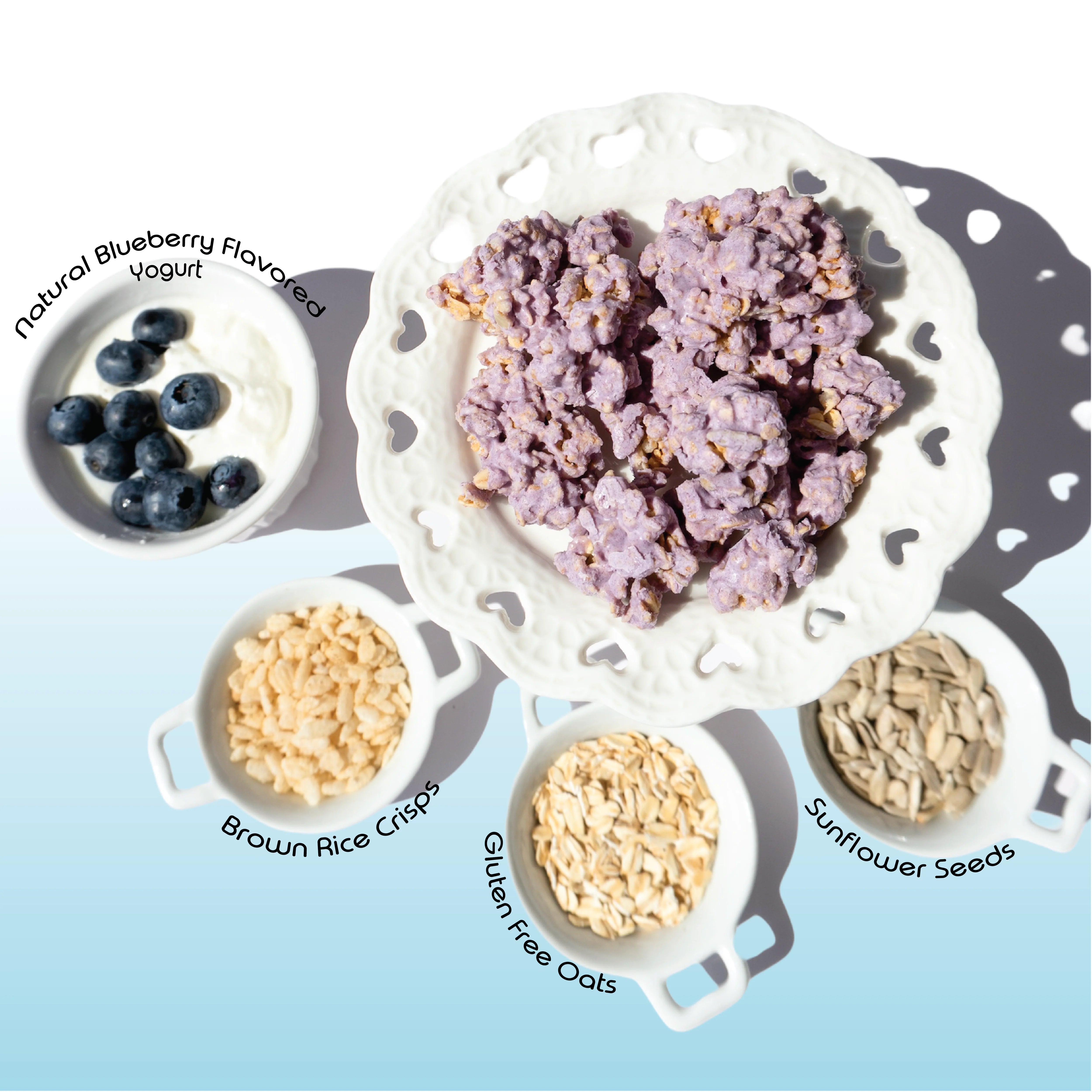 Happy Munch Blueberry Bliss yogurt clusters on a white plate with bowls of blueberries, brown rice crisps, gluten-free oats, and sunflower seeds.
