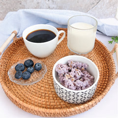 Cup of coffee, glass of milk, bowl of Happy Munch Blueberry Bliss, and blueberries on a woven tray.