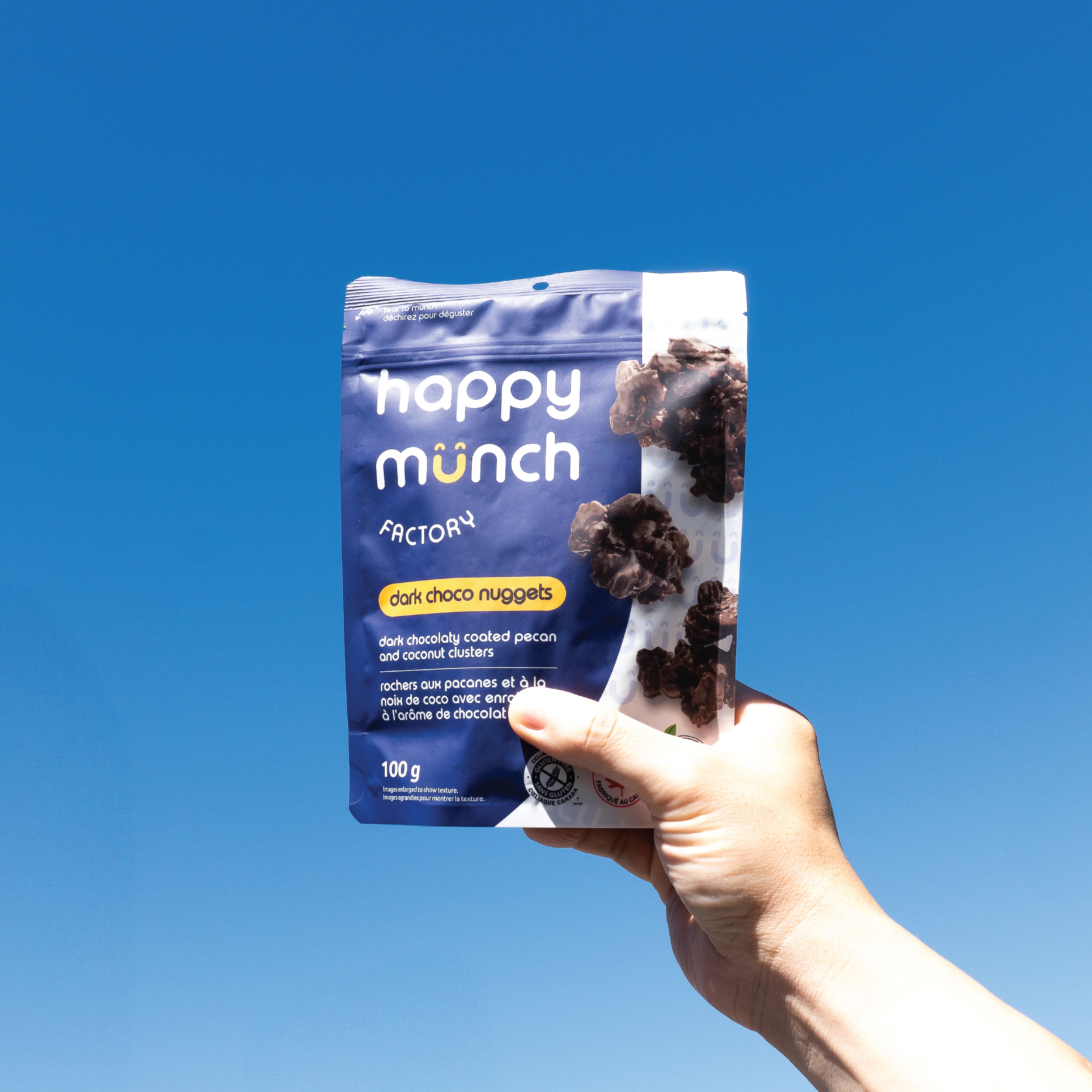 Hand holding a package of 'happy munch dark choco nuggets' against a clear blue sky.
