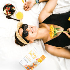 Woman lying on a white blanket with a sunflower, wearing sunglasses and a visor, next to a yogurt parfait and a Happy Munch Mango Mania pouch.
