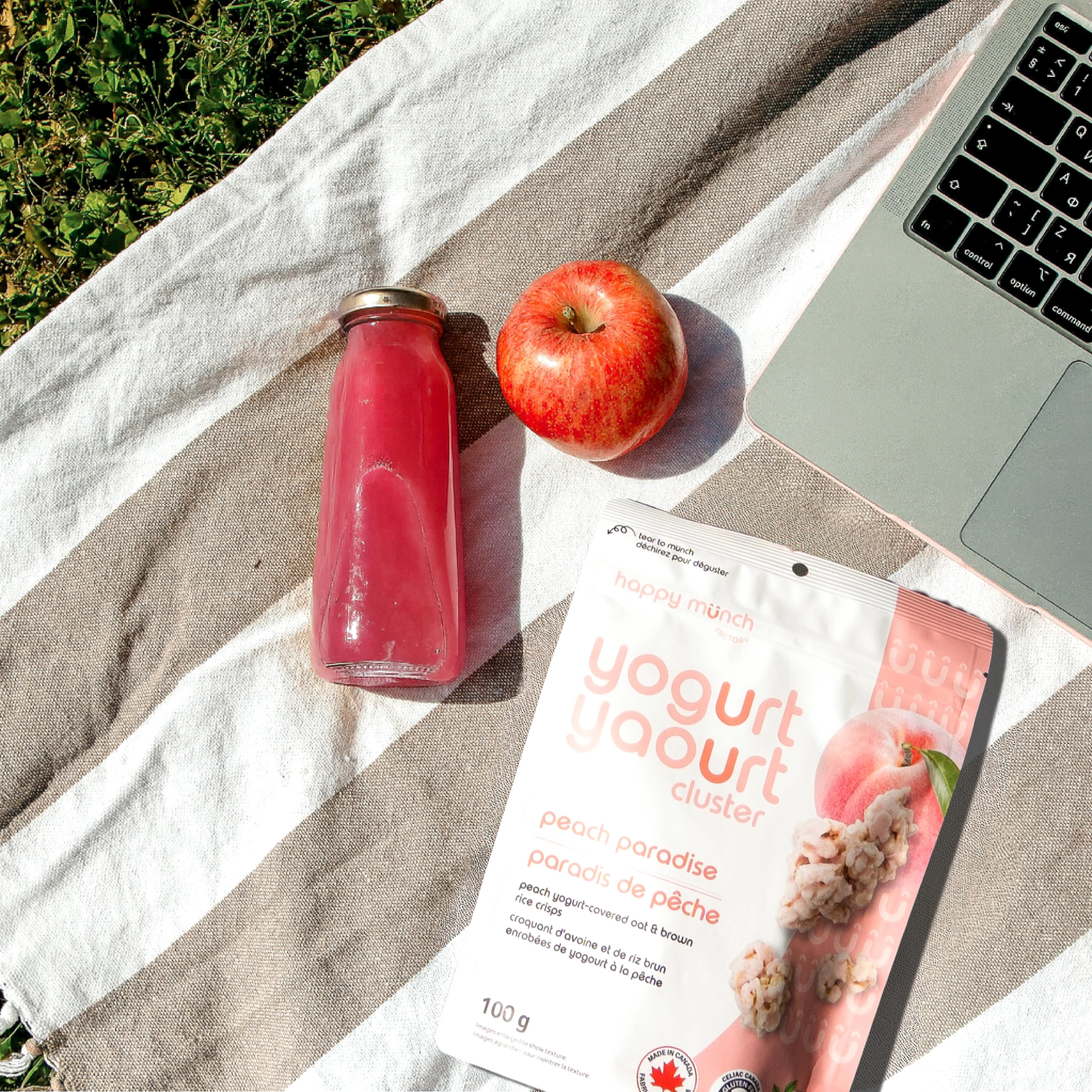 Happy Munch Peach Paradise Yogurt Clusters, red apple, pink bottle, and laptop on a striped towel outdoors.