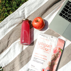 Happy Munch Peach Paradise Yogurt Clusters, red apple, pink bottle, and laptop on a striped towel outdoors.
