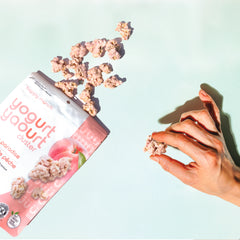 Hand holding a yogurt cluster snack next to Happy Munch Peach Paradise Yogurt Cluster Pouch on a light blue background